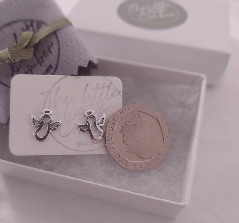 May include: Two silver angel earrings in a white box with a white card that says "The Little Angel Co.". A British one pound coin is next to the earrings for size comparison.