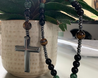 Può includere: Un rosario di perline con un ciondolo a forma di croce argentata. Le perline sono un mix di pietre nere, marroni, verdi e occhio di tigre. La croce ha un design semplice e lucido. Il rosario è drappeggiato su una pianta in vaso.