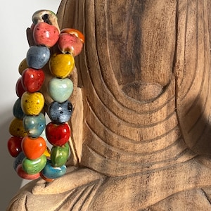 May include: A multi-strand bracelet featuring small, heart-shaped ceramic beads in a variety of vibrant colors including red, yellow, blue, orange, and green. The beads have a glossy, speckled finish and are strung together in a cluster.