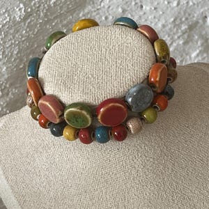 Può includere: Un braccialetto colorato con perline con un mix di perline ovali e rotonde in vari colori, tra cui rosso, verde, giallo, blu e arancione. Il braccialetto è esposto su una superficie di colore neutro.