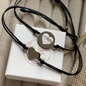 Könnte beinhalten: Zwei schwarze Armbänder aus Schnur mit silbernen Herzanhängern. Ein Anhänger ist ein volles Herz, der andere ein offenes Herz. Die Armbänder befinden sich in einer weißen Schachtel mit dem aufgedruckten Text "The Little Things In Other".