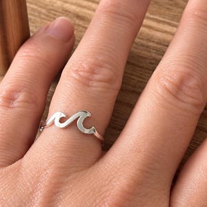 May include: A polished silver ring featuring a continuous wave design that wraps around the finger. The band is thin and smooth, showcasing a minimalist ocean-inspired motif with two distinct crests formed by the metal shape on the front.