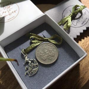 Puede incluir: Un colgante plateado con un surfista sobre una ola, presentado en una caja blanca con interior gris. Una moneda de cinco peniques y una cinta verde tambi&eacute;n est&aacute;n presentes. La caja tiene el texto "little earth amber JEWELLERY".