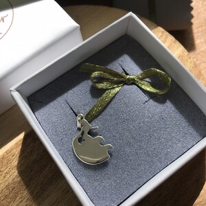 Puede incluir: Colgante de plata en una caja de regalo. El colgante presenta la silueta estilizada de una mujer con un vestido fluido. La caja es blanca con interior gris y una cinta verde atada con un lazo. El colgante se muestra sobre una superficie gris.