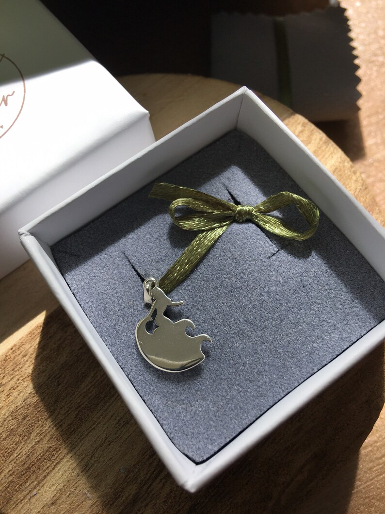 Puede incluir: Colgante de plata en una caja de regalo. El colgante presenta la silueta estilizada de una mujer con un vestido fluido. La caja es blanca con interior gris y una cinta verde atada con un lazo. El colgante se muestra sobre una superficie gris.