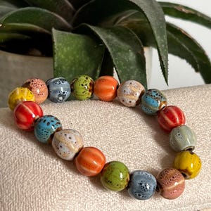May include: A bracelet made of ceramic beads in various colours, including yellow, red, blue, green, orange, and grey. The beads have speckled and striped patterns. The bracelet is displayed on a neutral-toned surface.