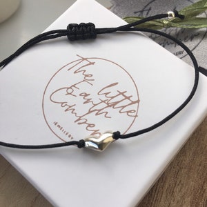 Op de afbeelding: Een zwart koordarmband met een zilveren hartbedel. Het armband is vastgemaakt met een knoop en heeft een zilveren kraal aan het einde. Het armband ligt op een witte doos met de tekst "The Little Earth Co. Jewellery" erop.