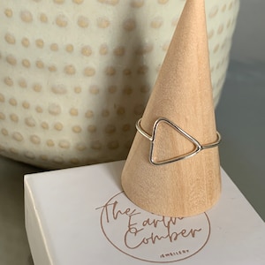 Könnte beinhalten: Ein silberner Dreiecksring auf einem hölzernen Ringständer. Der Ständer steht auf einer weißen Box mit dem aufgedruckten Text "The Earth & Comber Jewellery".