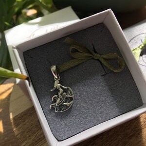 Puede incluir: Colgante plateado de una surfista en una ola, presentado en una caja de regalo blanca con interior gris y una cinta verde. El fondo muestra hojas verdes.