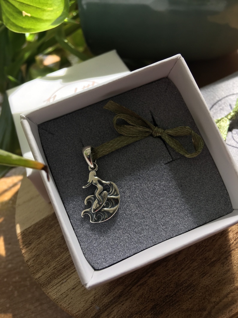 Puede incluir: Colgante plateado de una surfista en una ola, presentado en una caja de regalo blanca con interior gris y una cinta verde. El fondo muestra hojas verdes.