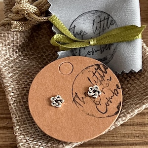 Può includere: Orecchini a bottone argentati a forma di nodo su un cartoncino rotondo marrone. Un piccolo panno grigio e un sacco di iuta sullo sfondo. Il cartoncino ha un logo stampato.