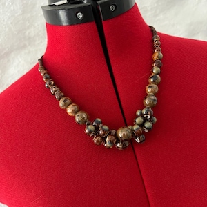 Könnte beinhalten: Eine Halskette mit braunen und goldfarbenen Perlen unterschiedlicher Größe. Die Halskette wird auf einer roten Schneiderpuppe präsentiert. Die Perlen sind in der Mitte geclustert und auf einer dunklen Schnur aufgefädelt.