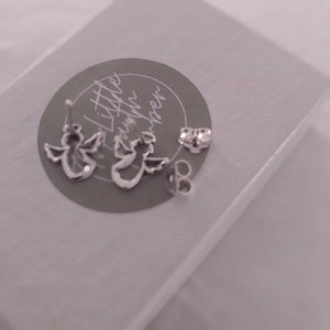 May include: A pair of silver angel wing earrings with a small silver post and back. The earrings are on a gray background with the text "Little Gatherings" written in cursive.