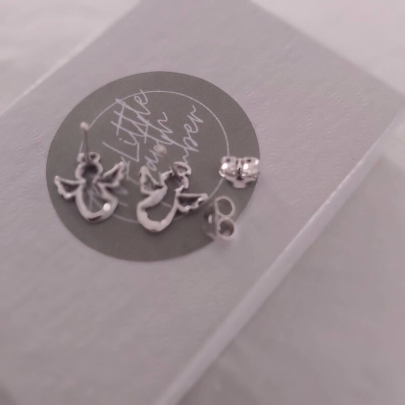 May include: A pair of silver angel wing earrings with a small silver post and back. The earrings are on a gray background with the text "Little Gatherings" written in cursive.