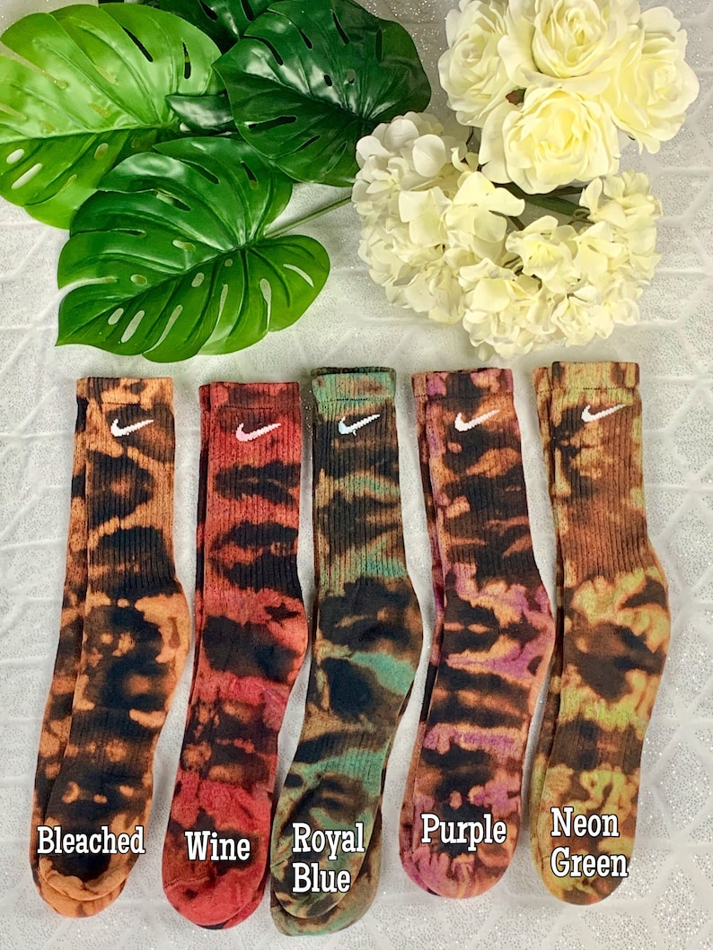 May include: Five pairs of crew socks in various colors, each with a tie-dye pattern and a white Nike swoosh logo. Colors include bleached, wine, royal blue, purple, and neon green. The socks are displayed on a white surface with green leaves and white flowers.