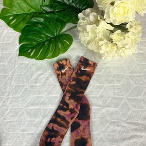 May include: A pair of tie-dye socks with a brown, black, and purple pattern. The socks feature the Nike swoosh logo in white. The socks are displayed with green leaves and white flowers.