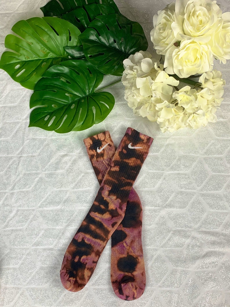 May include: A pair of tie-dye socks with a brown, black, and purple pattern. The socks feature the Nike swoosh logo in white. The socks are displayed with green leaves and white flowers.