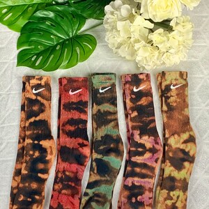 May include: Five pairs of crew socks with a tie-dye pattern in shades of orange, red, green, purple, and brown. Each sock features a white Nike swoosh logo. The socks are displayed on a white surface with green leaves and white flowers.