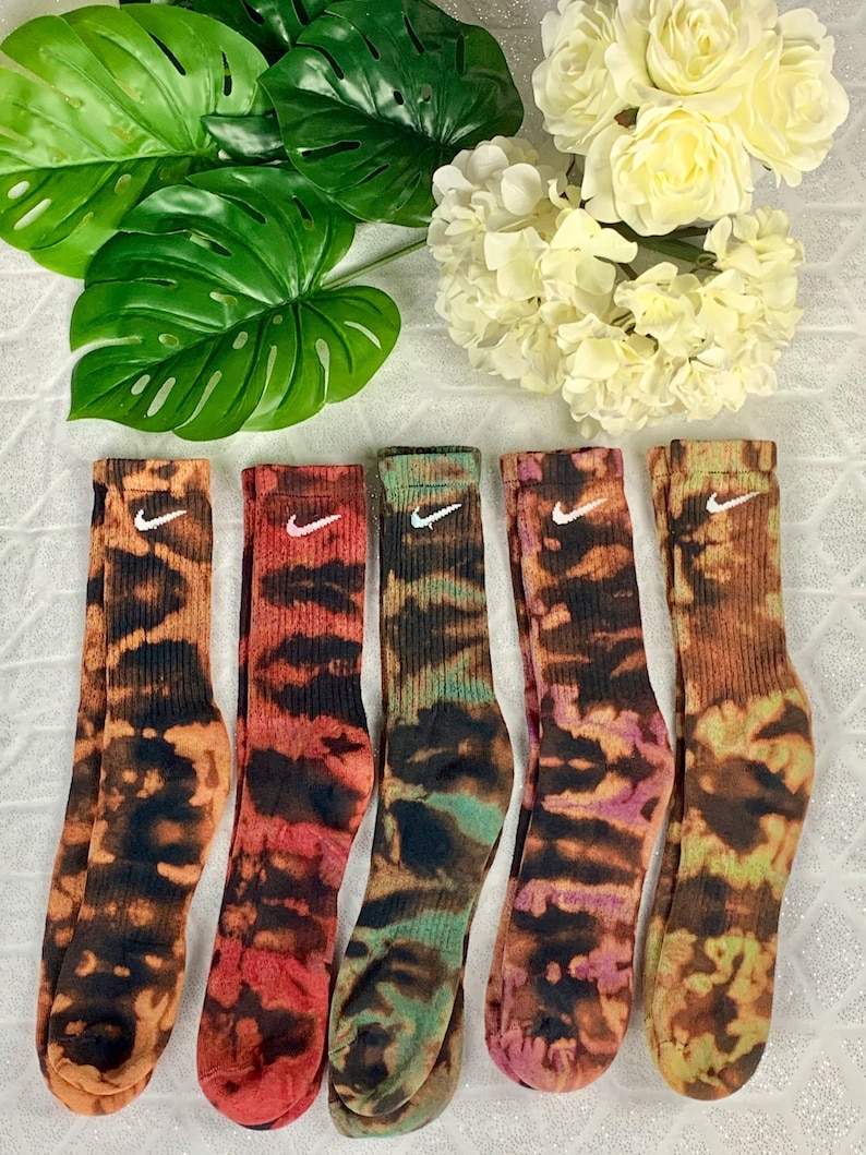 May include: Five pairs of crew socks with a tie-dye pattern in shades of orange, red, green, purple, and brown. Each sock features a white Nike swoosh logo. The socks are displayed on a white surface with green leaves and white flowers.