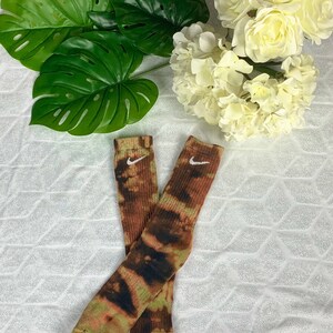 May include: A pair of tie-dye socks with a brown, green, and black pattern. The socks have a white Nike logo near the top. The socks are displayed on a white, textured surface with green leaves and white flowers in the background.