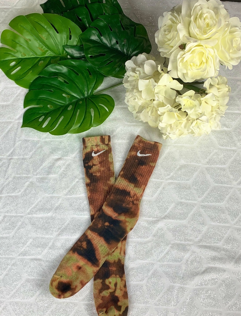 May include: A pair of tie-dye socks with a brown, green, and black pattern. The socks have a white Nike logo near the top. The socks are displayed on a white, textured surface with green leaves and white flowers in the background.