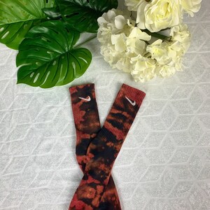 May include: A pair of tie-dye socks in shades of red, black, and brown. The socks feature the Nike swoosh logo in white near the top. The socks are displayed on a white surface with green leaves and white flowers in the background.