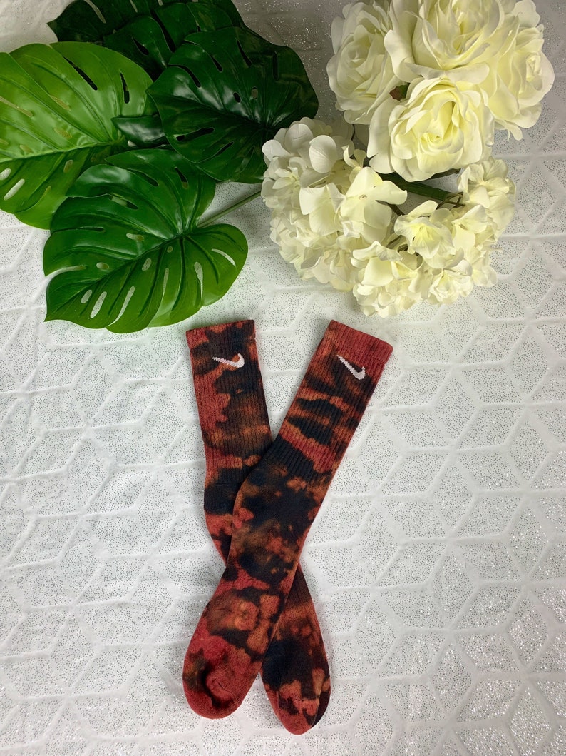May include: A pair of tie-dye socks in shades of red, black, and brown. The socks feature the Nike swoosh logo in white near the top. The socks are displayed on a white surface with green leaves and white flowers in the background.