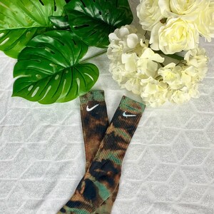 May include: Two pairs of crew socks with a tie-dye pattern in shades of brown, green, and black. Each sock features a white Nike swoosh logo near the top. The socks are displayed on a white, textured surface.