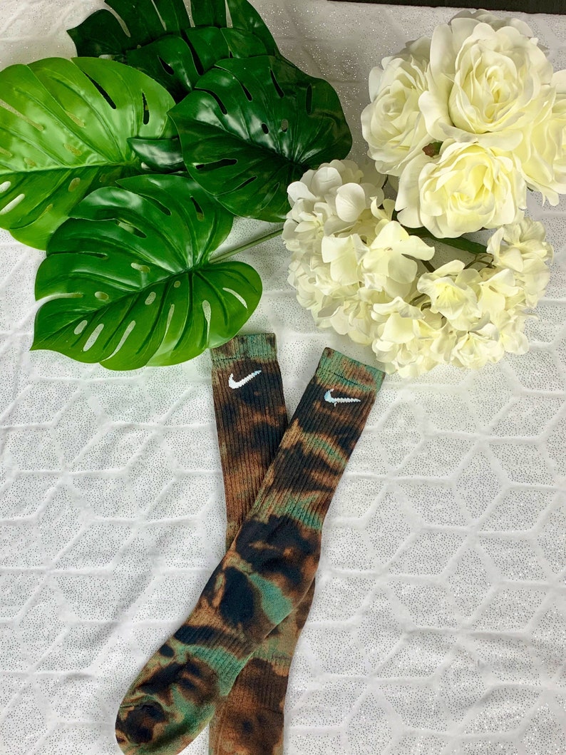 May include: Two pairs of crew socks with a tie-dye pattern in shades of brown, green, and black. Each sock features a white Nike swoosh logo near the top. The socks are displayed on a white, textured surface.