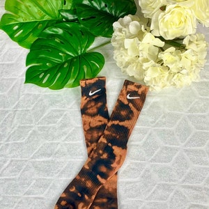 May include: A pair of crew socks with a tie-dye pattern in shades of brown and black. The socks feature the Nike swoosh logo in white. The socks are displayed on a white, textured surface, with green leaves and white flowers in the background.