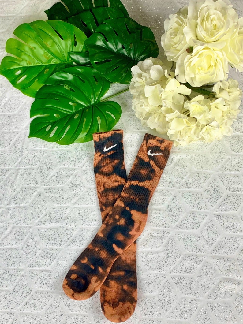 May include: A pair of crew socks with a tie-dye pattern in shades of brown and black. The socks feature the Nike swoosh logo in white. The socks are displayed on a white, textured surface, with green leaves and white flowers in the background.