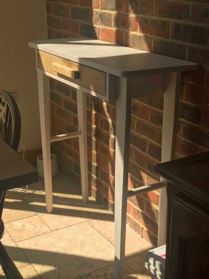 Op de afbeelding: Een kleine, wit en houtkleurige bijzettafel met een enkele lade. De tafel heeft een rechthoekig blad en vier slanke poten. De lade is van natuurlijk hout en heeft een eenvoudige, rechthoekige handgreep. De tafel is ongeveer 76 cm hoog.