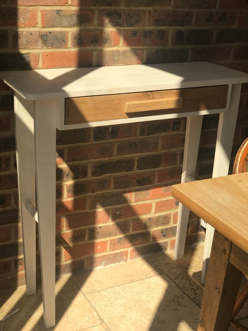 Op de afbeelding: Een wit geschilderde consoletafel met een enkele lade. De lade is gemaakt van natuurlijk hout en heeft een eenvoudige, rechthoekige handgreep. De tafel heeft een rechthoekig blad en vier rechte poten. De tafel is ongeveer 76 cm lang.