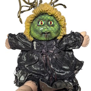 Pode incluir: Uma boneca personalizada com um rosto verde, olhos vermelhos e um penteado de serpentes semelhante ao da Medusa. A boneca usa um vestido de renda preta com uma saia com babados. A pele da boneca &eacute; de cor p&ecirc;ssego claro.