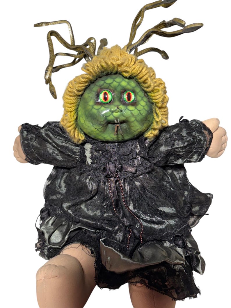 Pode incluir: Uma boneca personalizada com um rosto verde, olhos vermelhos e um penteado de serpentes semelhante ao da Medusa. A boneca usa um vestido de renda preta com uma saia com babados. A pele da boneca &eacute; de cor p&ecirc;ssego claro.