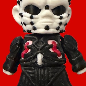 May include: A 3D-printed figurine of a character with a white head covered in black pins and large black eye sockets. The figure wears a black outfit with red accents and stands against a red background.