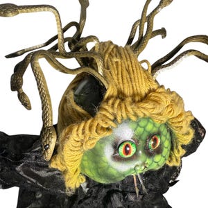Pode incluir: Uma boneca com o tema Medusa com um rosto verde, olhos vermelhos e verdes e um penteado de fio amarelo. A boneca tem cabelo de cobra cor de bronze e est&aacute; vestindo uma roupa de tecido preto.