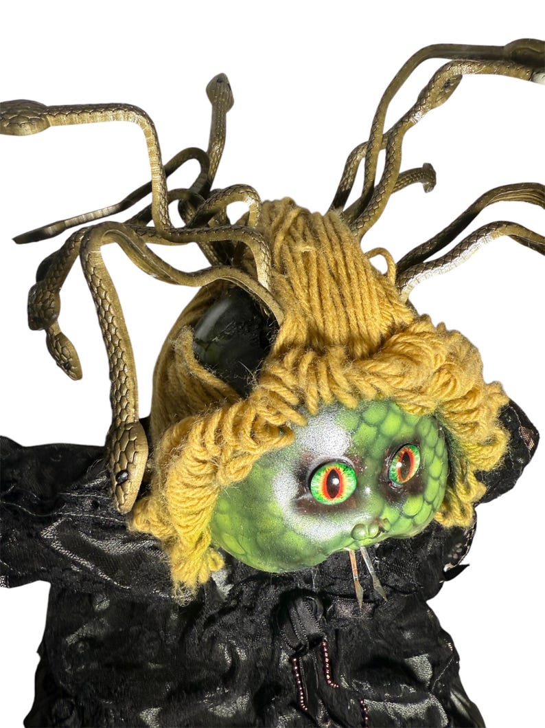 Pode incluir: Uma boneca com o tema Medusa com um rosto verde, olhos vermelhos e verdes e um penteado de fio amarelo. A boneca tem cabelo de cobra cor de bronze e est&aacute; vestindo uma roupa de tecido preto.