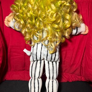 Pu&ograve; includere: Bambola fatta a mano con capelli ricci gialli e verdi, indossa un completo a righe bianche e nere. Le gambe e i piedi della bambola sono visibili, con scarpe nere. La bambola &egrave; sdraiata su una superficie rossa.