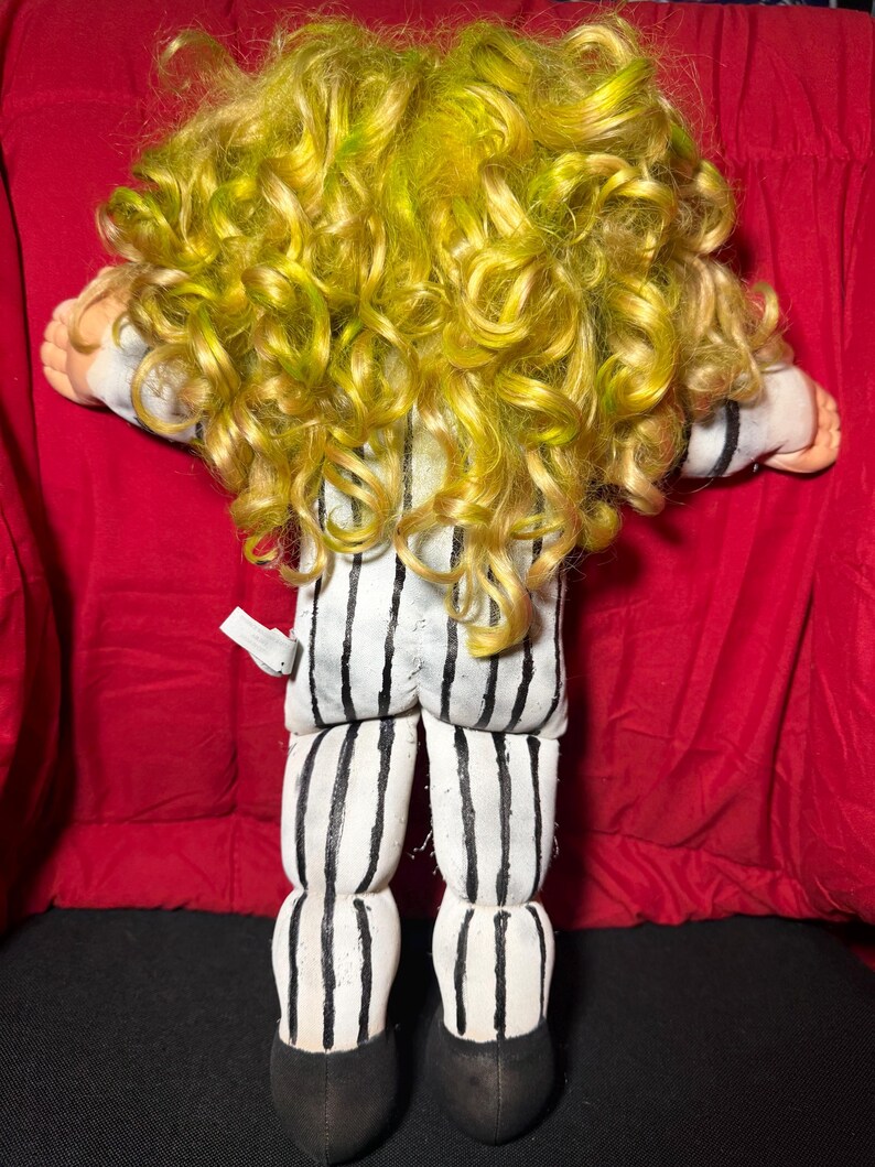 Pu&ograve; includere: Bambola fatta a mano con capelli ricci gialli e verdi, indossa un completo a righe bianche e nere. Le gambe e i piedi della bambola sono visibili, con scarpe nere. La bambola &egrave; sdraiata su una superficie rossa.