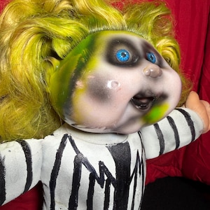 Peut inclure: Poupée avec un visage vert et noir, des yeux bleus et des cheveux jaunes. La poupée porte une tenue rayée noire et blanche. Le visage de la poupée a une expression peinte avec une bouche ouverte.