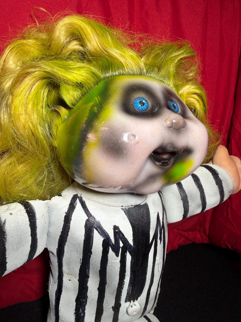 Pu&ograve; includere: Bambola con viso verde e nero, occhi blu e capelli gialli. La bambola indossa un completo a righe bianche e nere. Il viso della bambola ha un'espressione dipinta con la bocca aperta.