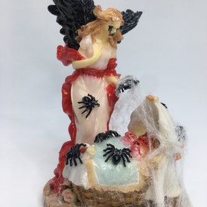 May include: A Halloween-themed figurine of an angel with black wings and a red sash. The angel is standing over a basket with spiders and a web. The basket contains small teddy bear heads and red objects.