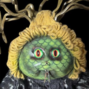 May include: A Medusa doll with a green, scaled face, yellow yarn hair, and metal snake tendrils. The doll has large, red and green eyes and a forked metal tongue. The doll is wearing a black, textured garment.