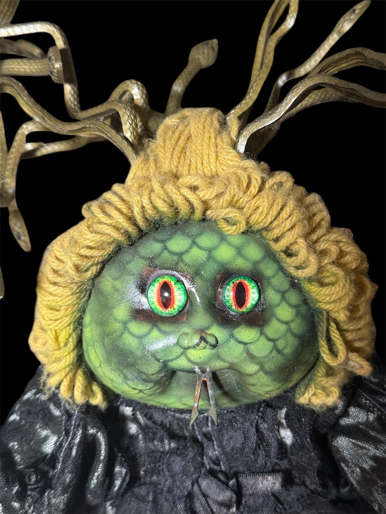 Pode incluir: Uma boneca com o tema Medusa com um rosto verde e escamoso, cabelo de fio amarelo e gavinhas de metal em forma de cobra. A boneca tem olhos grandes, vermelhos e verdes e uma l&iacute;ngua met&aacute;lica bifurcada. A boneca est&aacute; vestindo uma roupa preta texturizada.