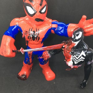 May include: A red and blue Spider-Man action figure with a black spider emblem on the chest, holding a sword that is piercing a black and white Venom action figure. The figures are on a black surface.