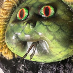 Pode incluir: Uma m&aacute;scara grotesca de pele verde com olhos grandes com pupilas vermelhas e pretas. A m&aacute;scara tem uma apar&ecirc;ncia de cobra, com uma juba amarela semelhante a fios e uma gola de renda preta. Uma pe&ccedil;a de metal se projeta da boca.