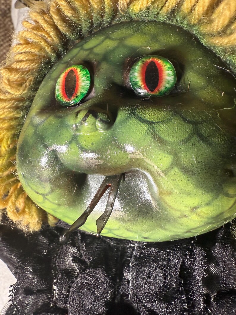 Pode incluir: Uma m&aacute;scara grotesca de pele verde com olhos grandes com pupilas vermelhas e pretas. A m&aacute;scara tem uma apar&ecirc;ncia de cobra, com uma juba amarela semelhante a fios e uma gola de renda preta. Uma pe&ccedil;a de metal se projeta da boca.