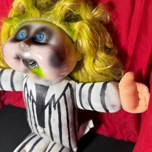 Pu&ograve; includere: Bambola di peluche con viso pallido, occhi blu e capelli gialli. La bambola indossa un abito a righe bianche e nere. Il viso della bambola ha trucco nero e muco verde che esce dalla bocca.