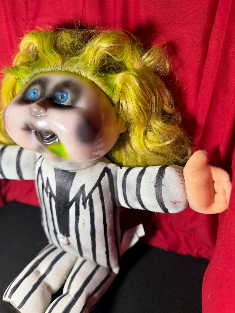 Pu&ograve; includere: Bambola di peluche con viso pallido, occhi blu e capelli gialli. La bambola indossa un abito a righe bianche e nere. Il viso della bambola ha trucco nero e muco verde che esce dalla bocca.